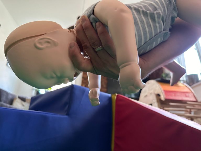 Eine Hand hält eine Puppe in einer schrägen Position über eine weiche Unterlage.