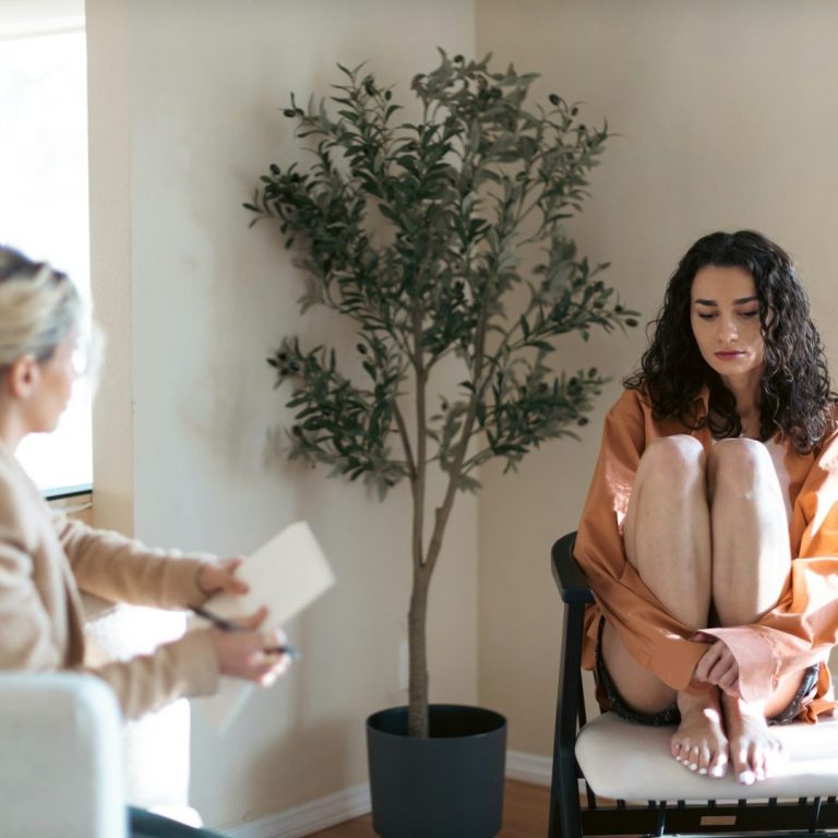 Betrieblicher Ersthelfer für psychische Gesundheit Eine sitzende Frau mit lockigem Haar in einem orangefarbenen Kleid neben einem Topfbaum.