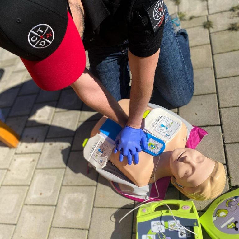Betriebliche Erste Hilfe mit Consulting + Training Dörfler Person führt CPR an einer Übungspuppe im Freien durch. Defibrillator ist nebenan.