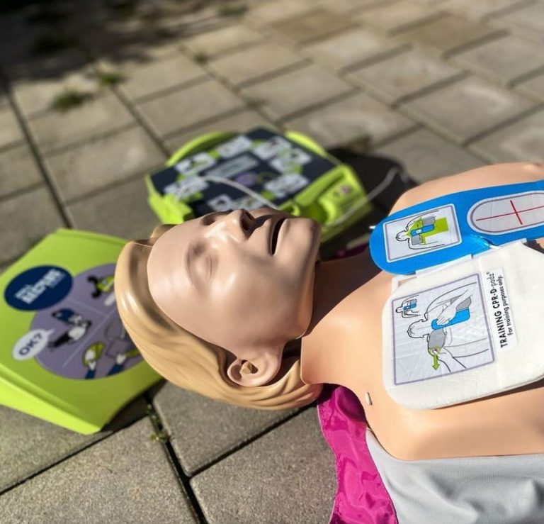 Ein Übungspuppe für Erste Hilfe mit Anleitungen auf der Brust, umgeben von Defibrillatoren.