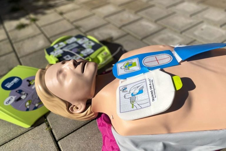 Übungspuppe für Herz-Lungen-Wiederbelebung mit Defibrillatoren im Hintergrund.