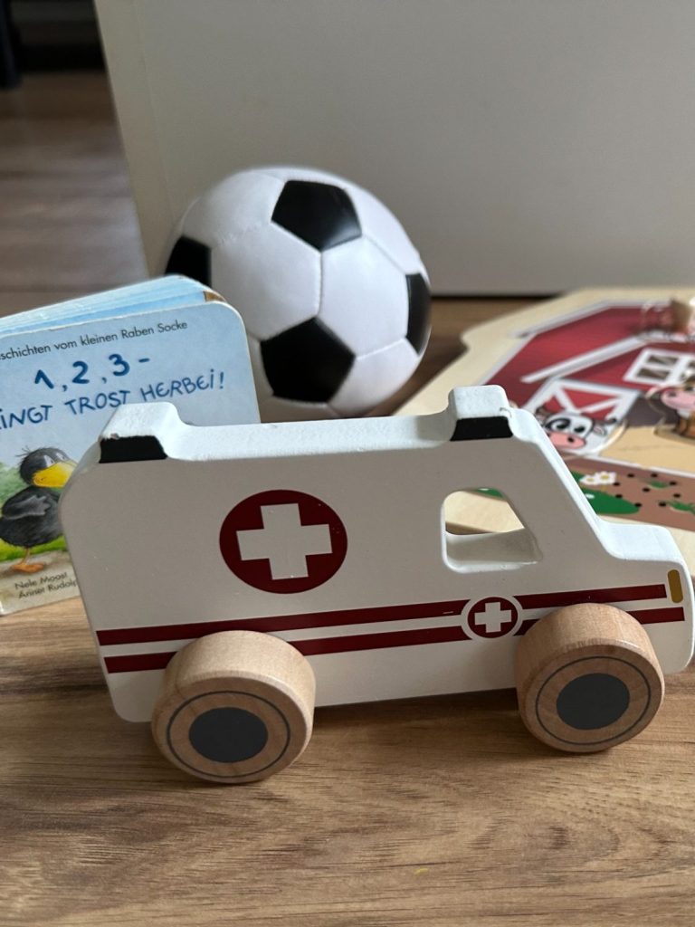 Holzambulanz mit roten Kreuz, umgeben von einem Fußball und Spielzeugbüchern.