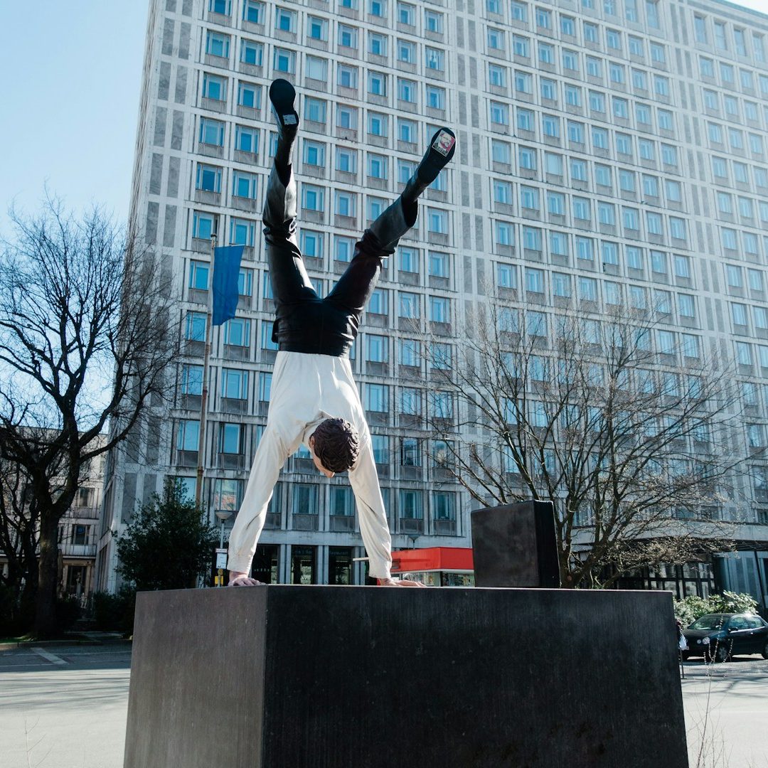 Führungskräfteschulungen Person in Hemd zeigt einen Handstand auf einem Podest vor einem modernen Gebäude.