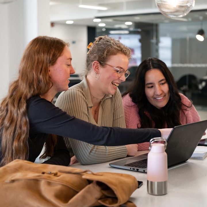 Gesündere Teams durch BGM Drei junge Frauen sitzen zusammen und schauen auf einen Laptop, lächeln und diskutieren.