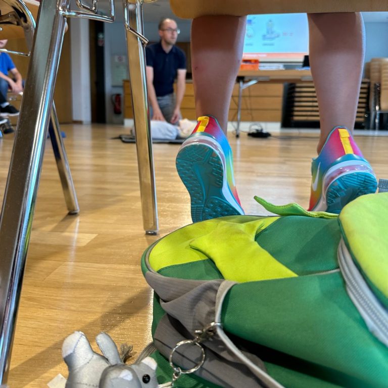 Beine einer Person in bunten Sportschuhen, im Hintergrund eine CPR-Übung.